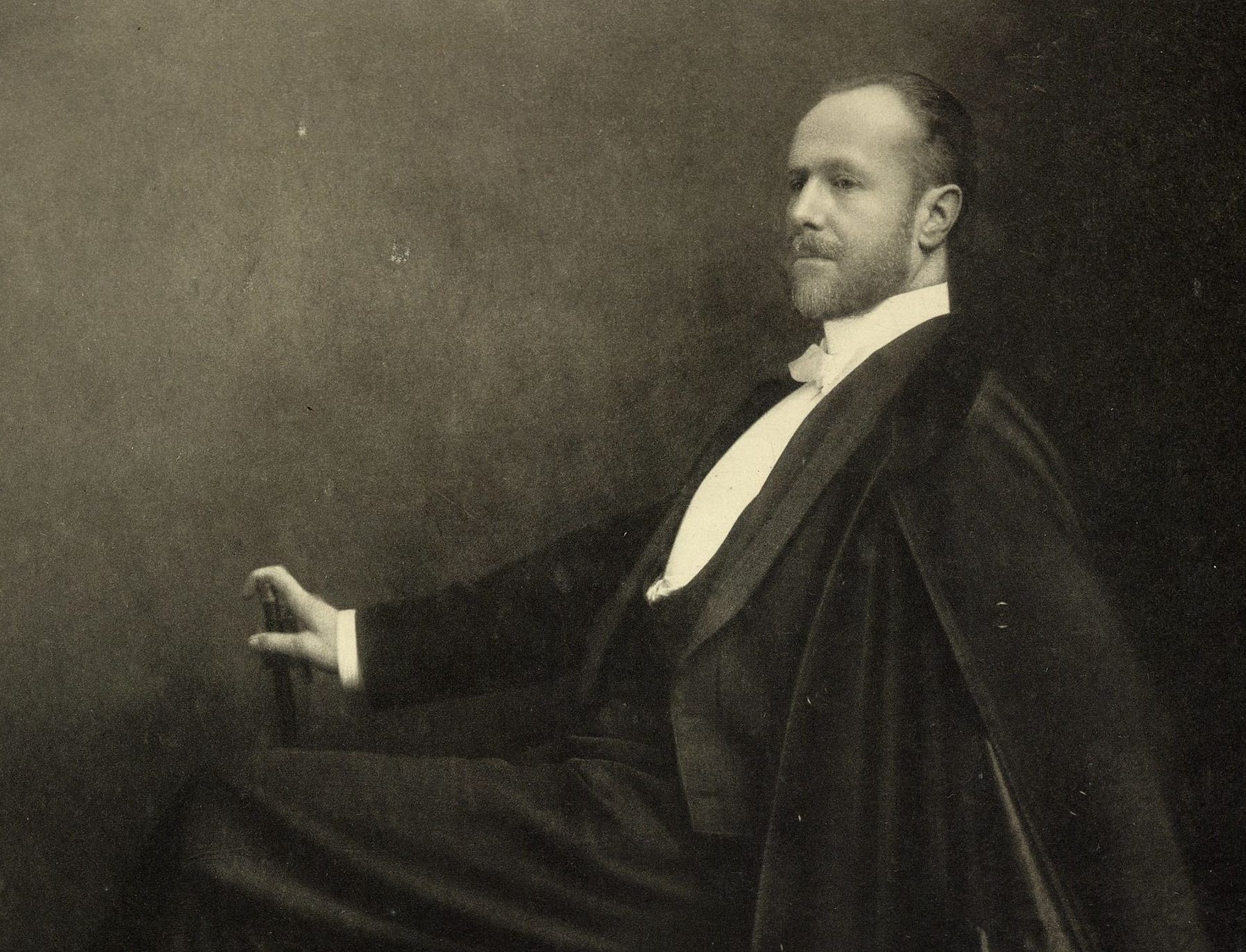 Black and white photograph of Fernand Khnopff, the Belgian artist and central figure of the Symbolist movement, posed seated in profile with his hand on a chair back, exuding a contemplative demeanor.